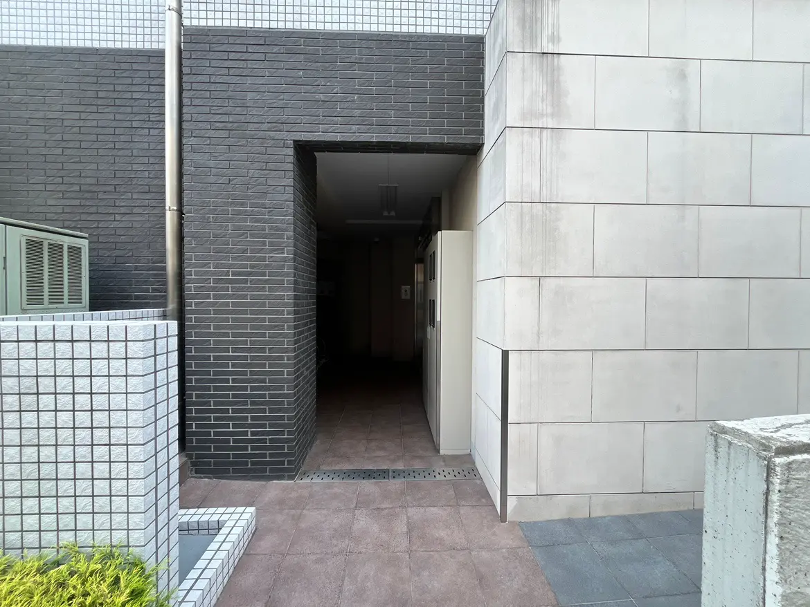 - モダンな建物の入口、黒いタイルとコンクリートの壁、左側にエアコンユニット、明るいタイルの床の風景