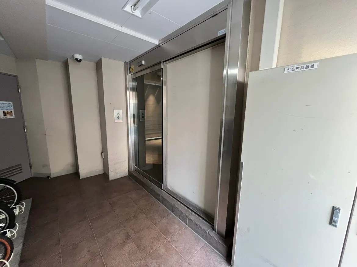 - エレベーターの前の風景、壁に設置された監視カメラと呼び鈴、左側には自転車が収納されている空間