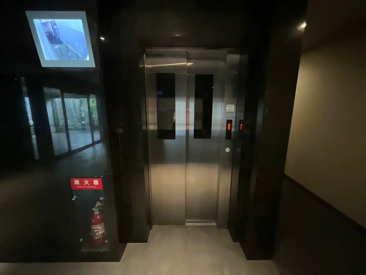 - モダンなデザインのエレベーターの入り口、壁面に設置された監視カメラと消火器の表示がある暗いトーンの廊下