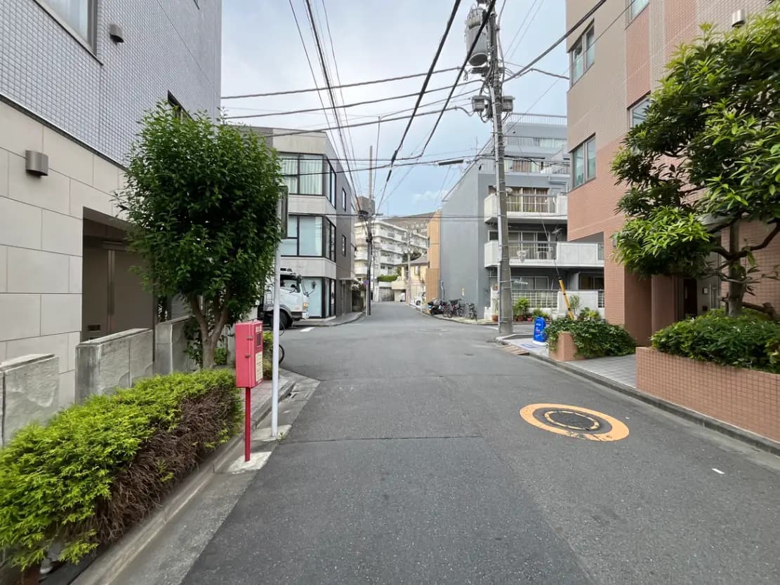 - 都市の静かな裏道、建物の間に並ぶ緑の植栽とピンクの郵便ポスト、晴れた空の風景