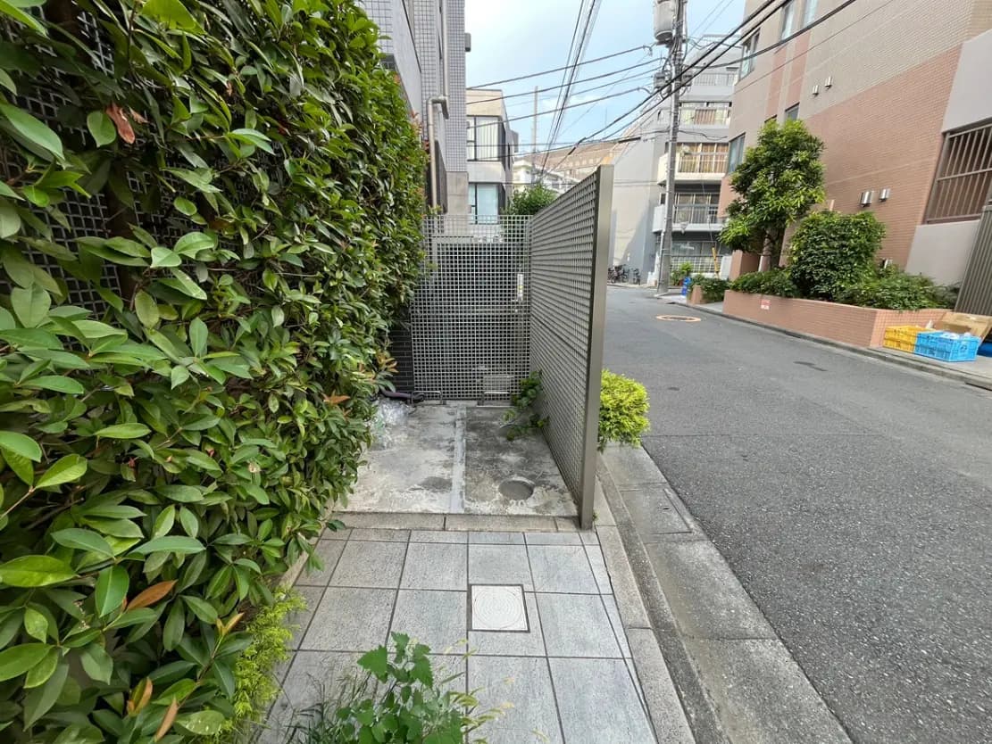 - 緑豊かな生垣とコンクリートの歩道がある静かな街角、右手には幹線道路と住宅が見える風景