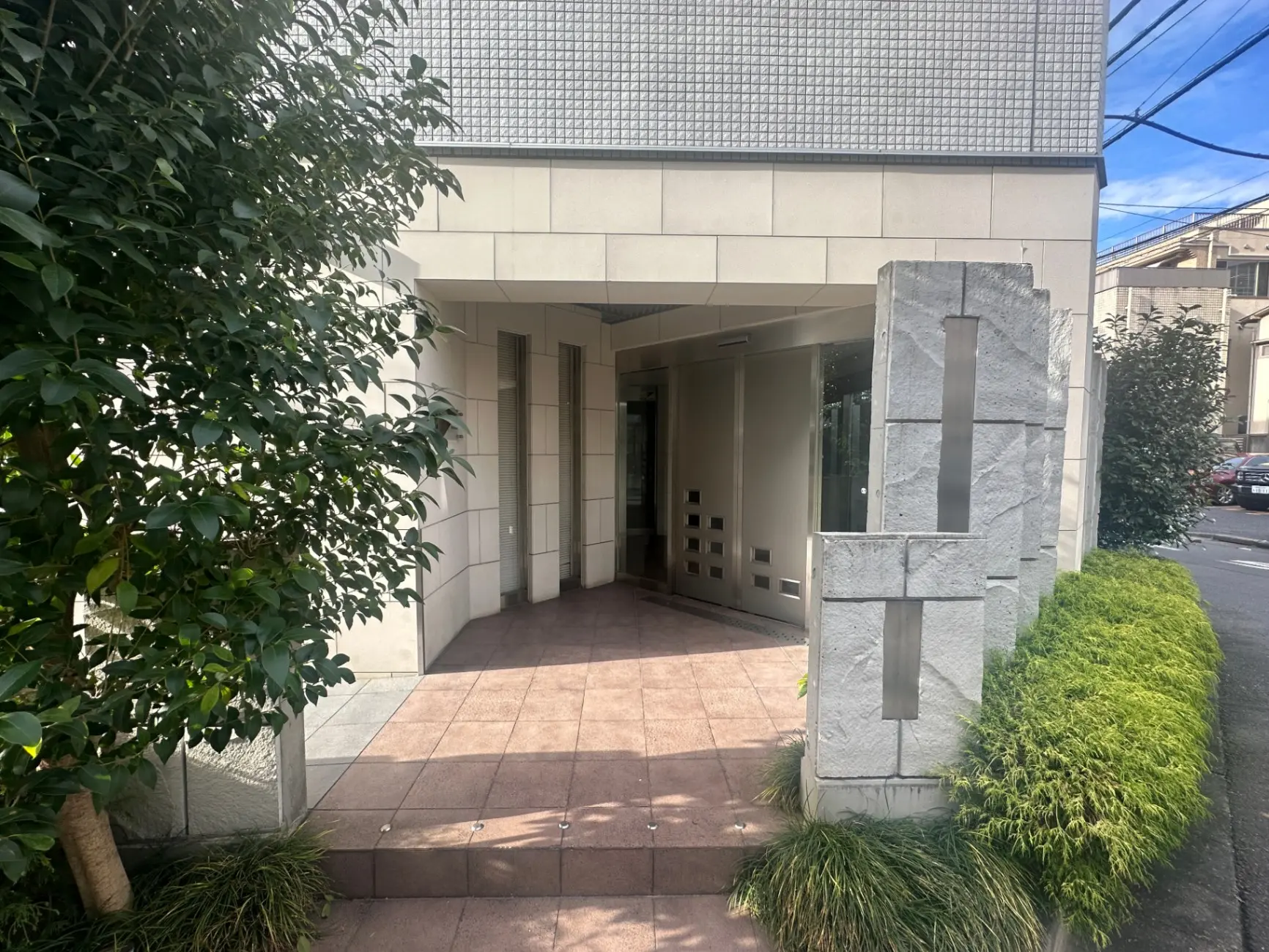 現代的な建物の入り口、石造りの柱とガラスドア、緑豊かな植栽がある外観