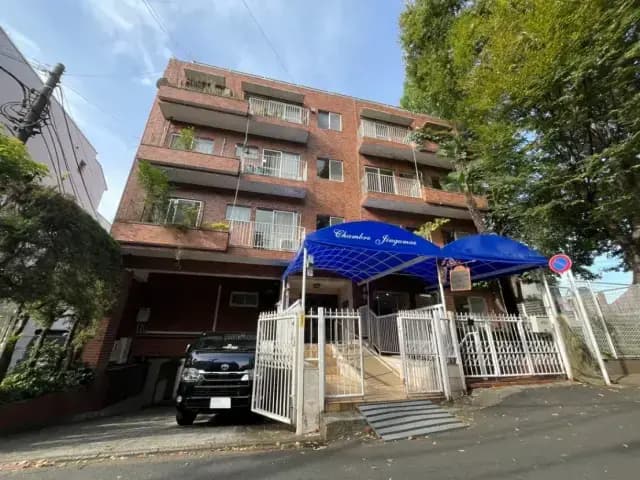 - レンガ造りのマンション、青いひさしが特徴的で、周囲に緑が感じられるエントランスの外観