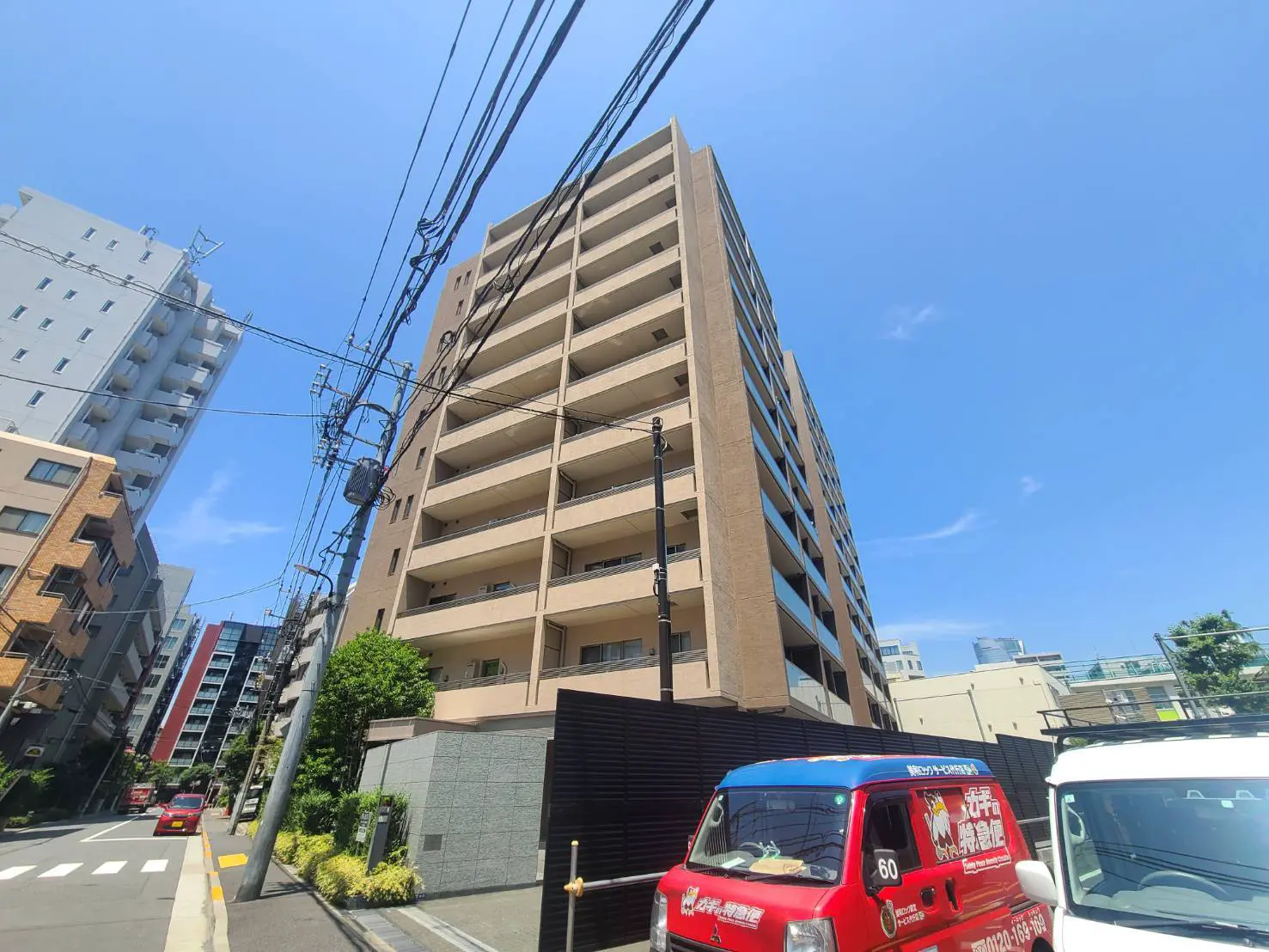 - 高層マンションの外観、青空を背景にしたモダンな建物と近隣の住宅地、電柱と電線が見える街並み