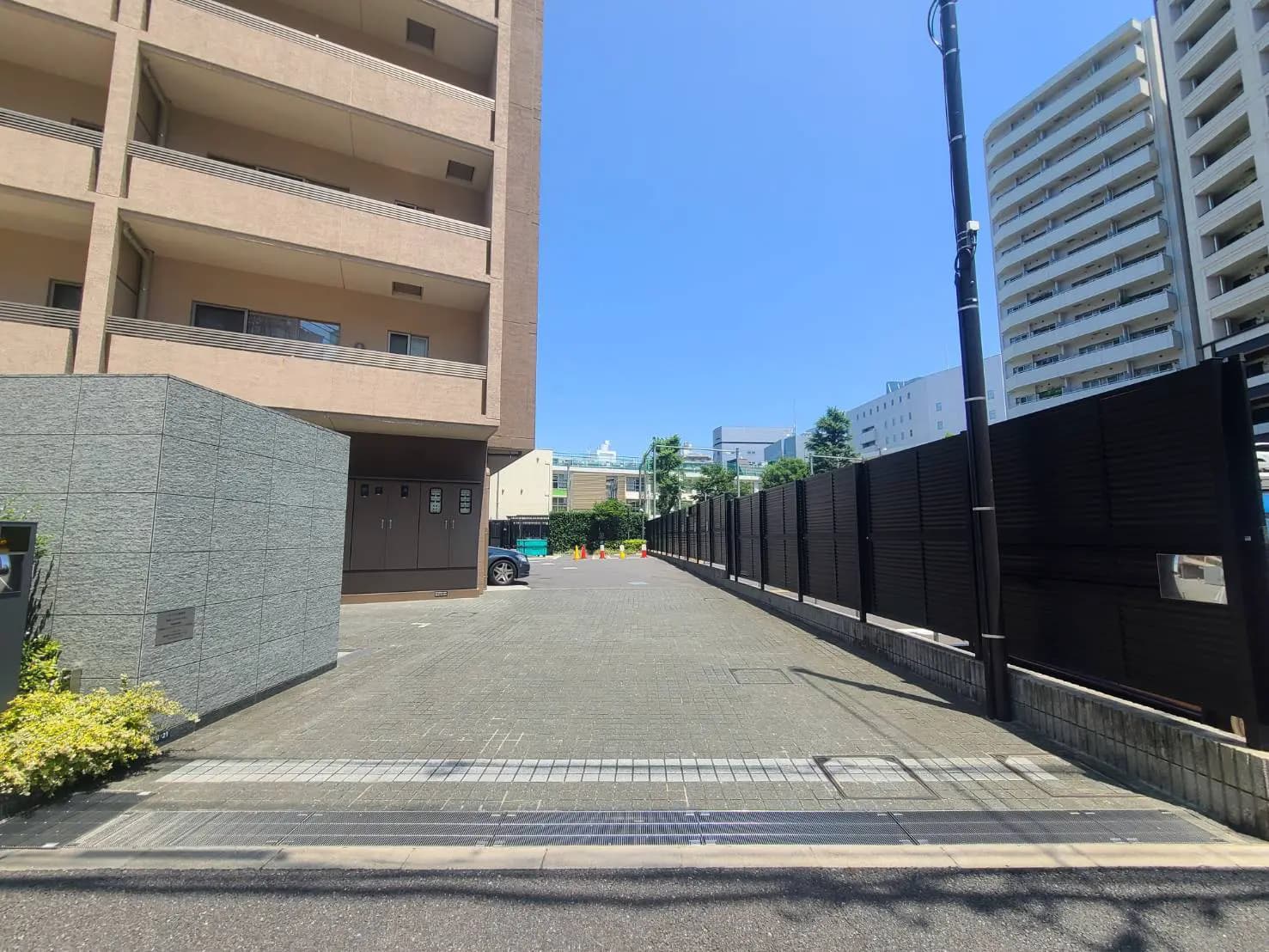 - 明るい青空の下、整備された駐車場と黒いフェンスで囲まれたアパートのエントランス周辺の風景