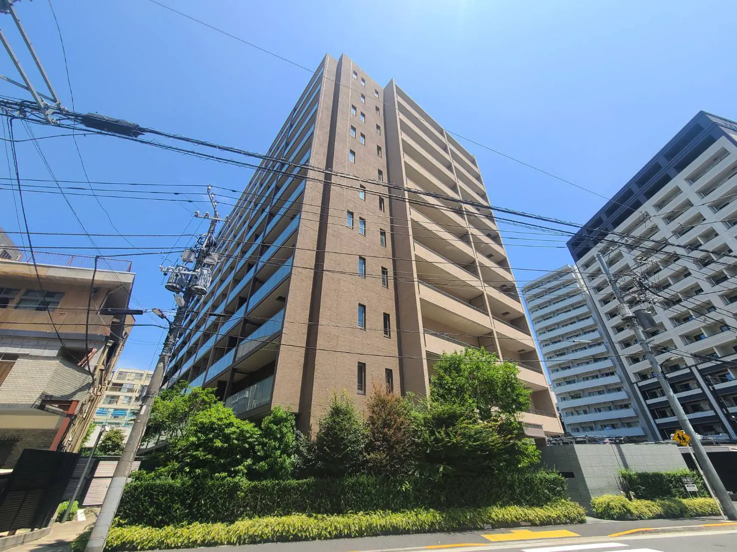 - 青空を背景にした現代的な外観のマンション、周囲に緑が多く、電線が交差する都市風景
