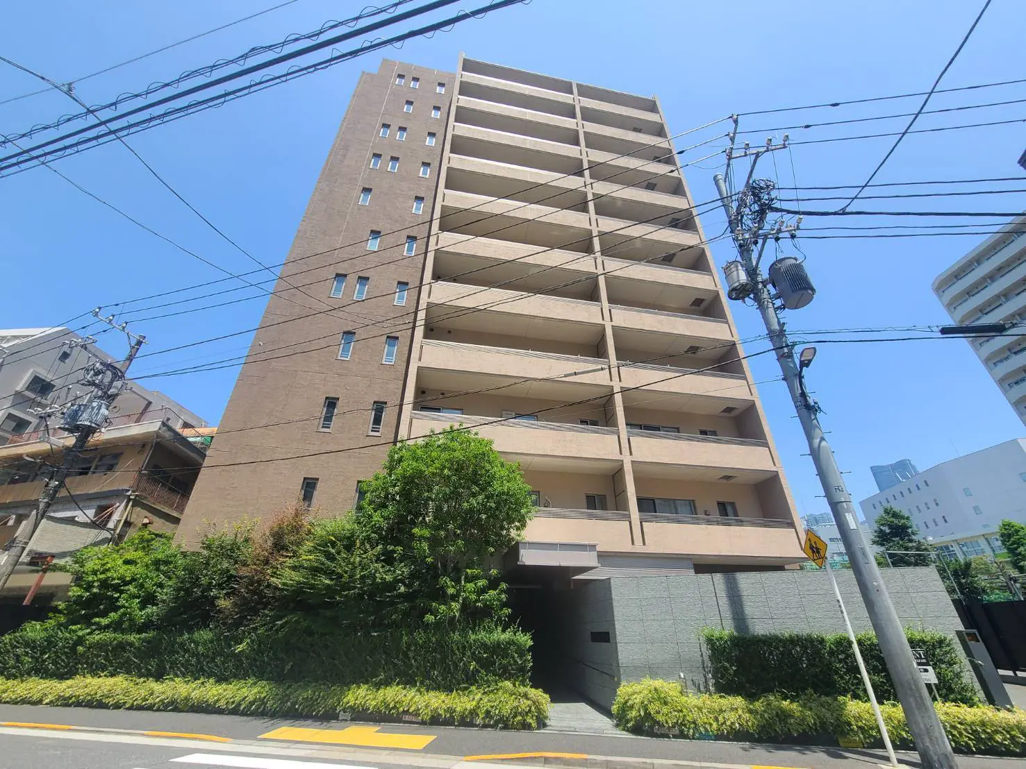 - ビルの外観、晴れた青空の下に立つ8階建てのマンション、周囲には緑豊かな植栽と電柱が見えるシーン