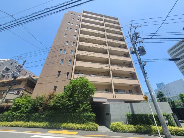 - ビルの外観、晴れた青空の下に立つ8階建てのマンション、周囲には緑豊かな植栽と電柱が見えるシーン