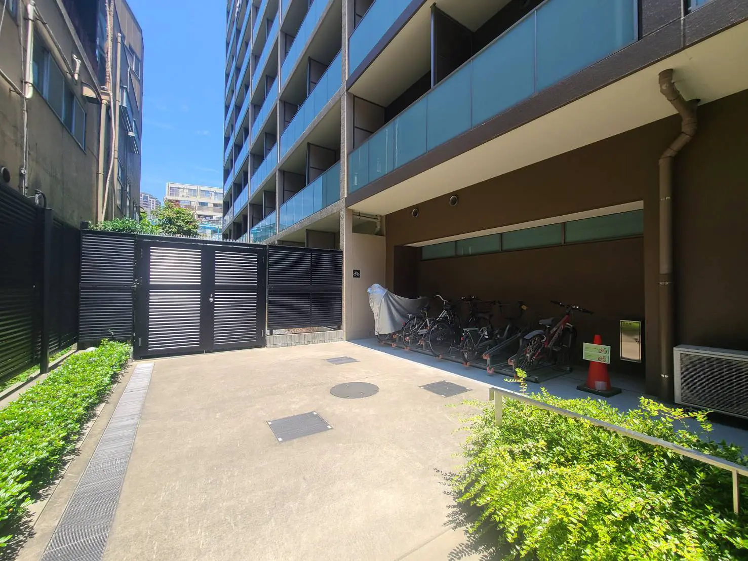 - 自転車置き場のある現代的なマンションの外観、手前には緑の植栽とシャッターゲートがある風景