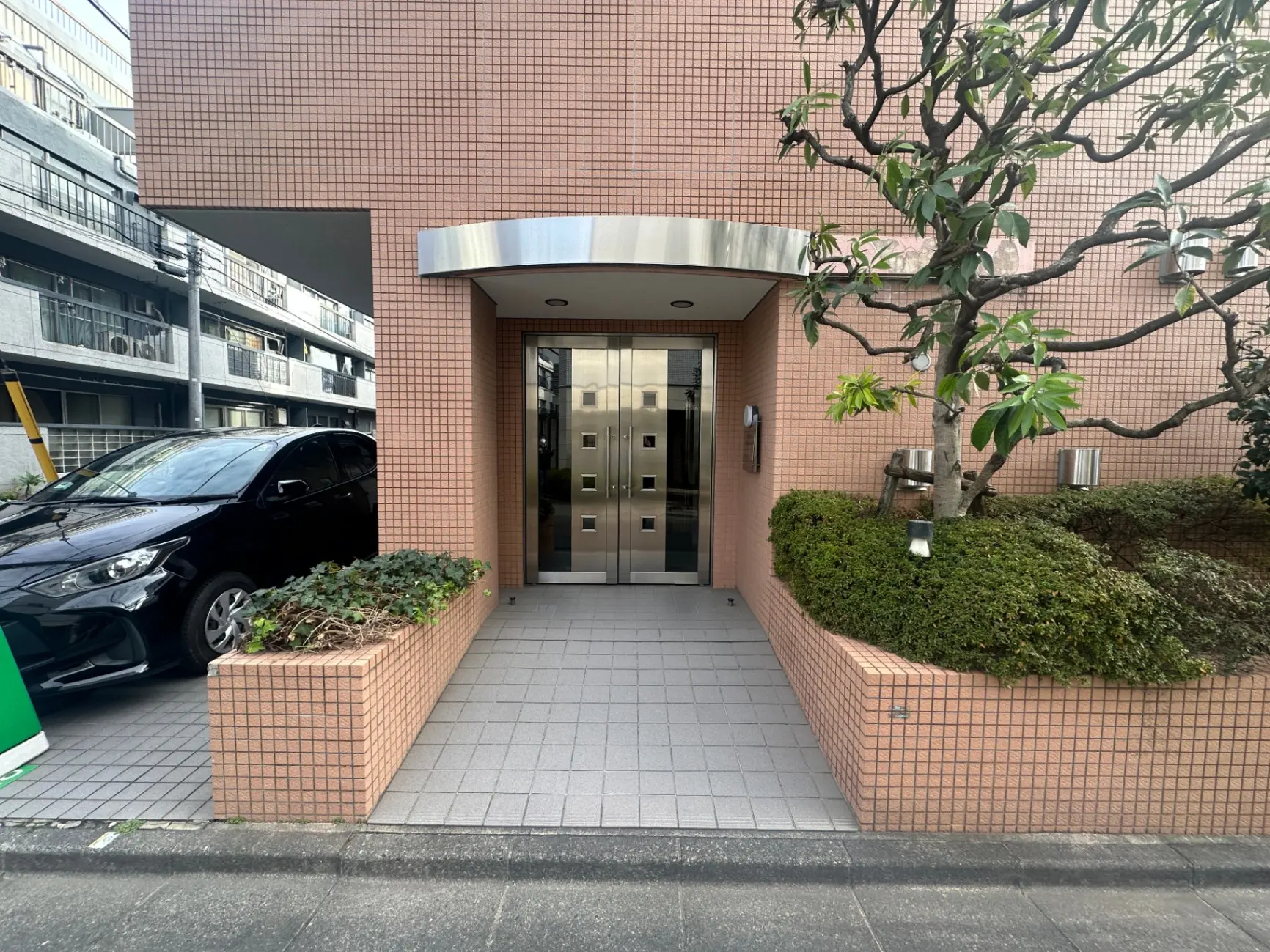 ピンクタイルの外壁が特徴的なアパートのエントランス、緑豊かな植木とクリーンな歩道