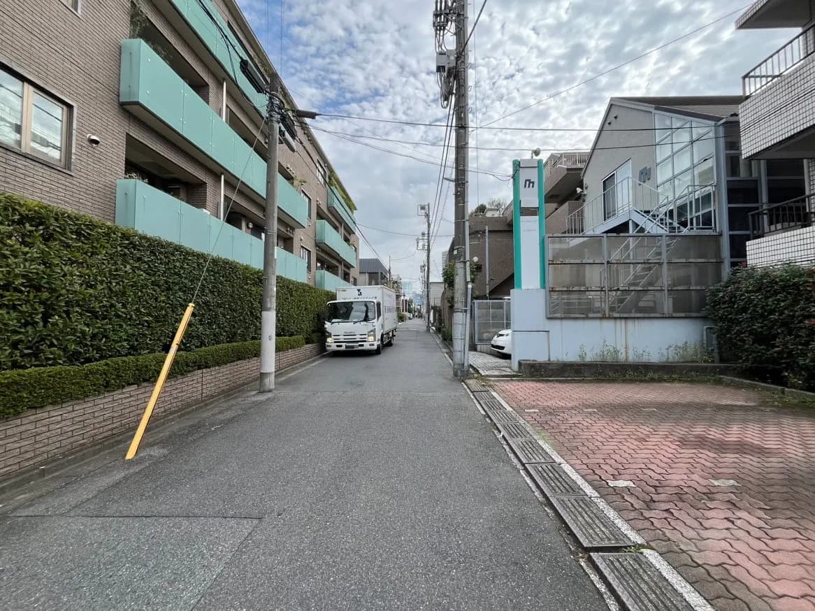 日本の住宅街の通り、左側に緑の低層アパート、右側には駐車場と白い個人宅
