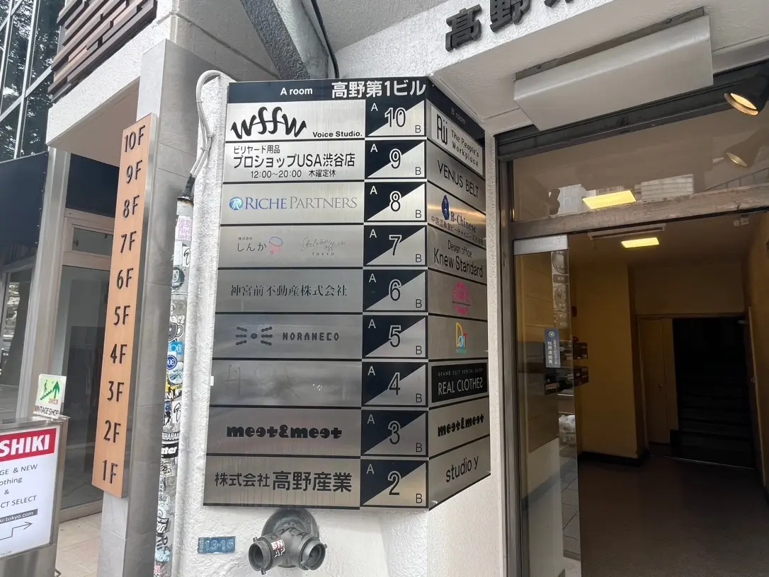 ビル入口の案内板、複数企業のロゴと階数表示、歩行者用道に面した建物入り口