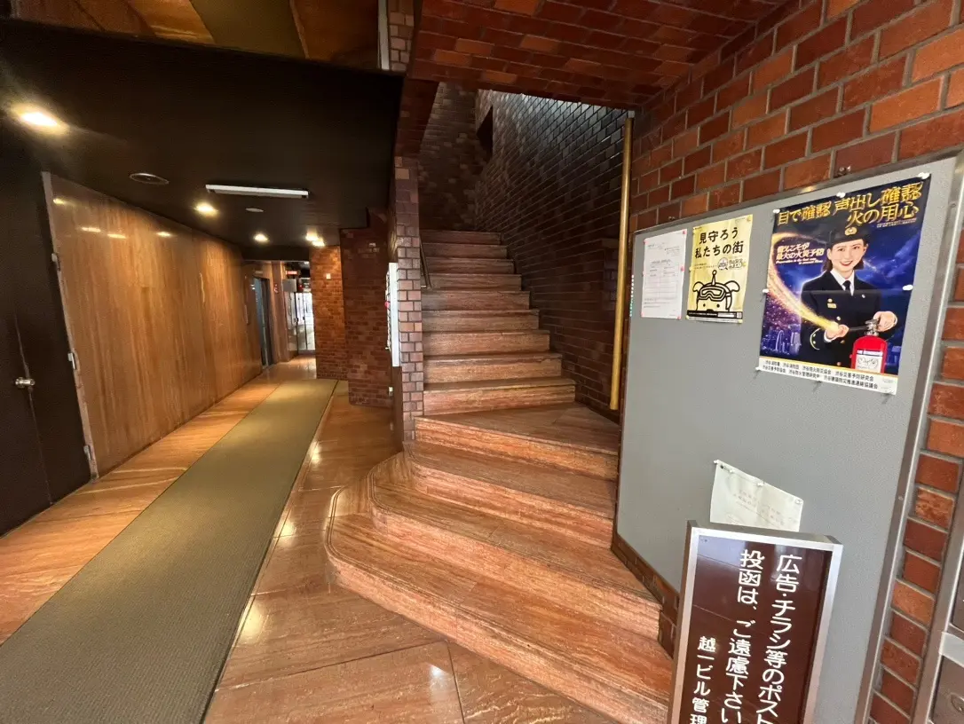 レンガ壁と木の手すりが特徴的な建物内の階段、ポスターや案内掲示が見られる廊下のインテリア