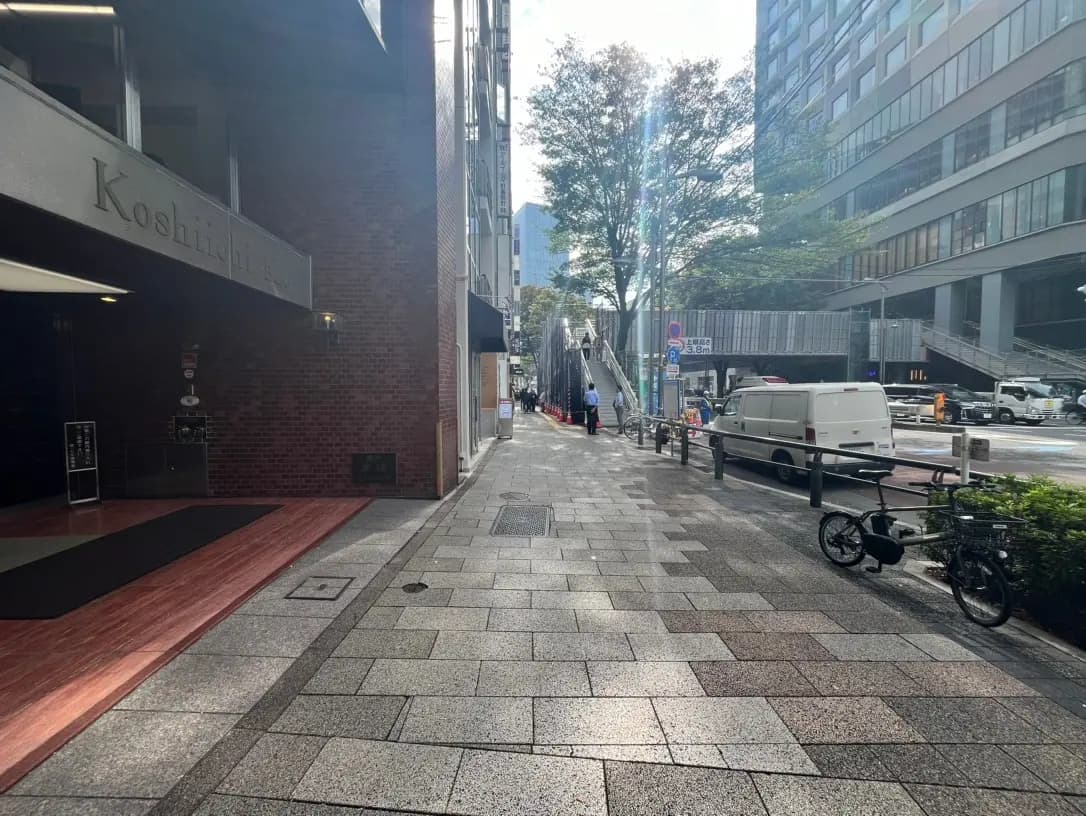 日当たりの良い市街地の歩道、レンガ造りの建物入口と自転車、遠くに歩行者と走行中の車が見える風景
