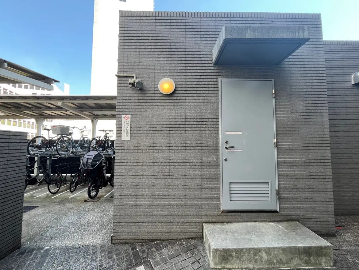 - 自転車置き場の入口、グレーの壁にドアと照明、奥に自転車のラックが見える都市の風景