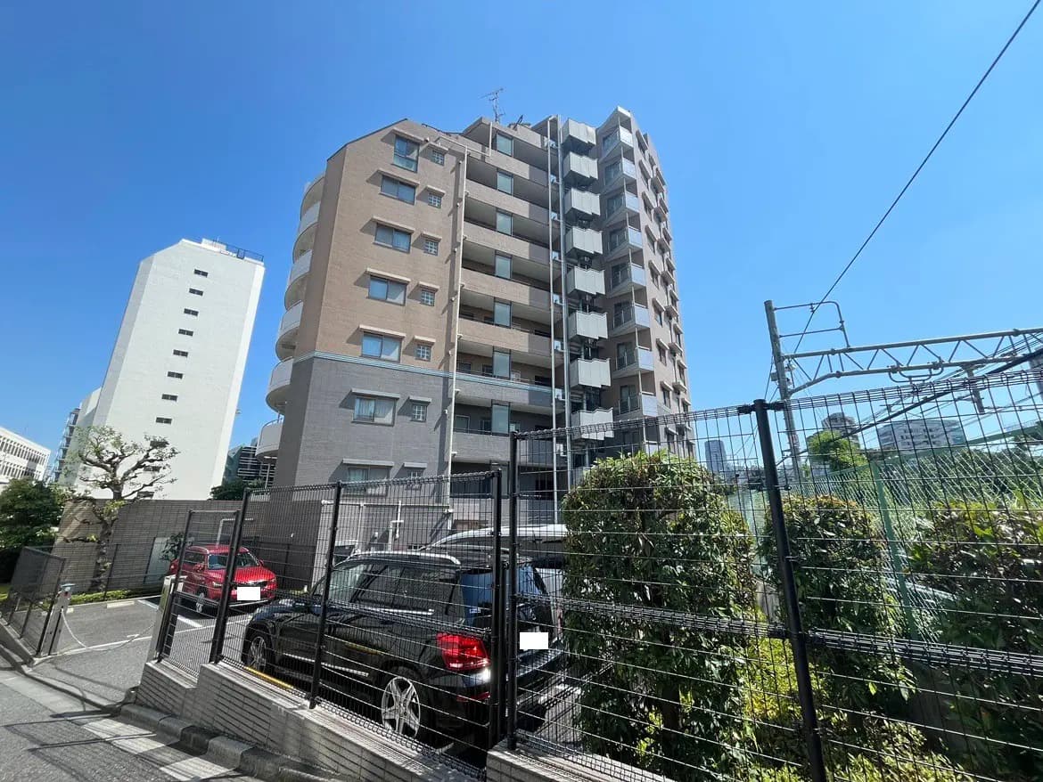 - 明るい青空の下に立つ高層マンション、周囲に駐車場と緑の植栽が見える都市の風景