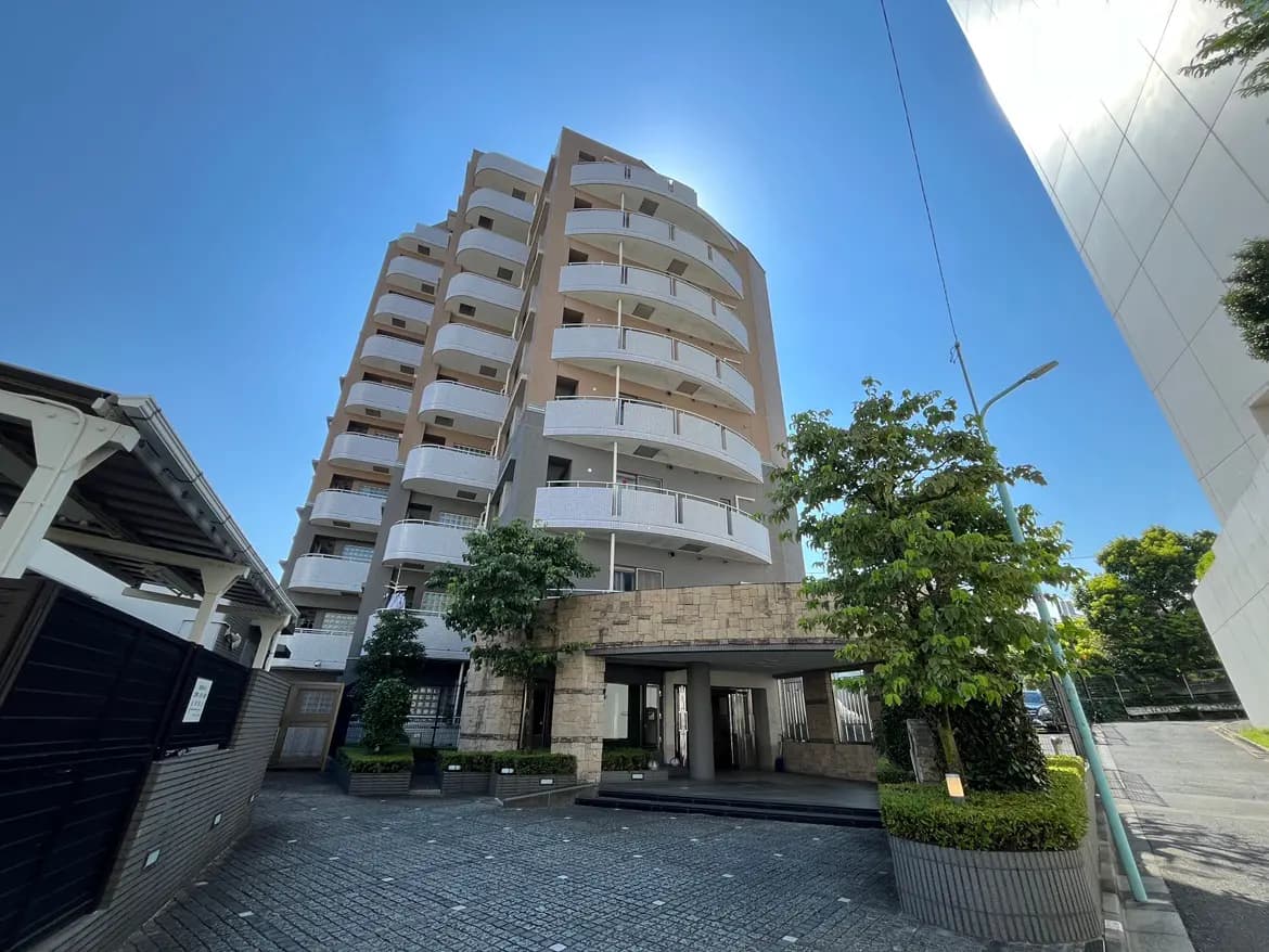 - 現代的なデザインの集合住宅、周囲に植栽が施されたエントランスと広い駐車場、青空を背景にした外観