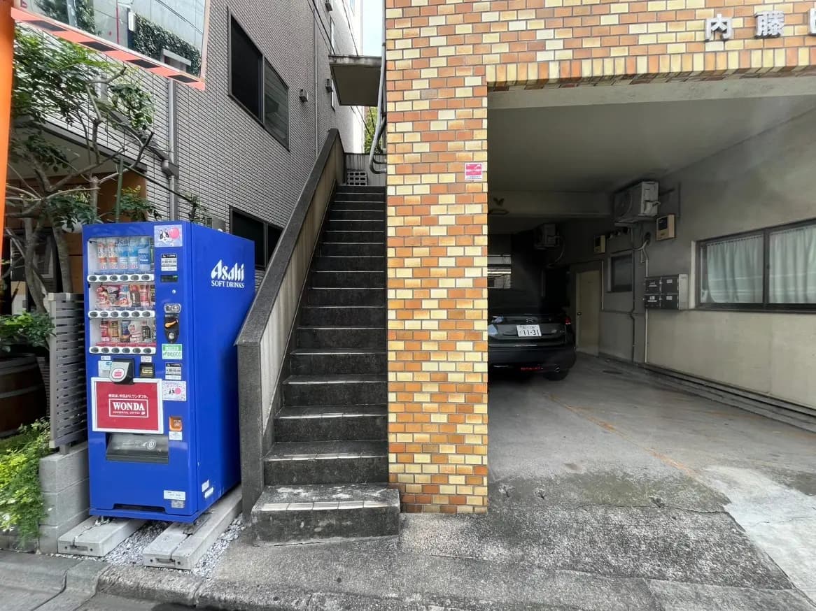 都市のアパート外観、階段と自動販売機、背景に駐車スペース