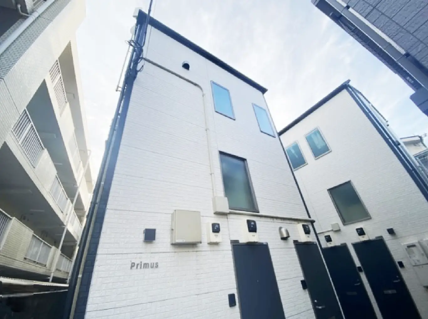 - 白い外壁の建物と隣接するアパート、明るい空の下での外観、Primusの名称が見えるエントランス。