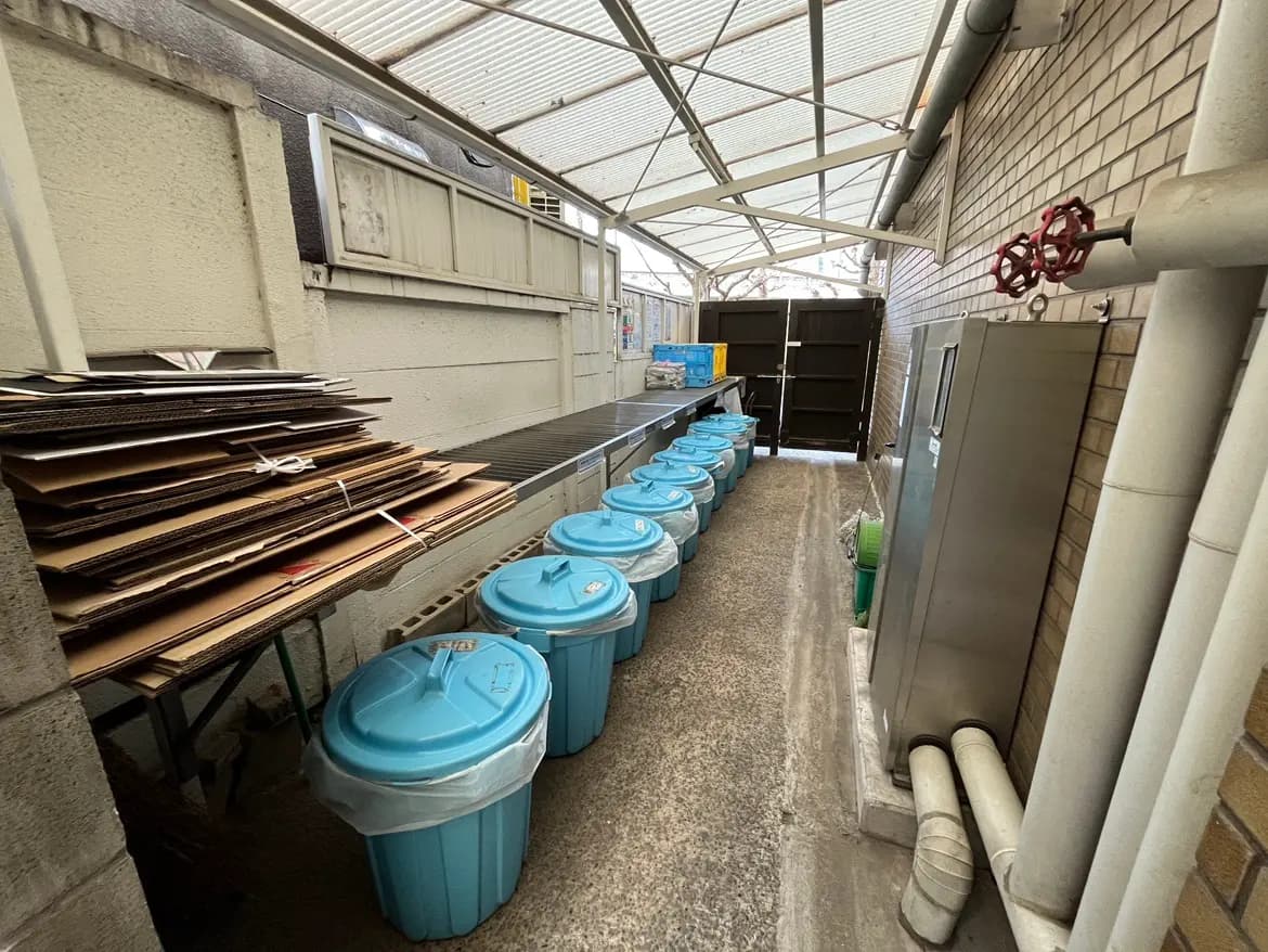 - 青いゴミ箱が並ぶ屋外の廊下、段ボールが積まれた台やパイプが見える清掃エリアの風景