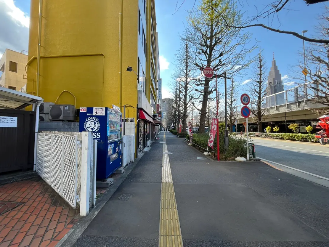 - 黄色い建物と自販機が並ぶ街道、青空と並木道が広がる都市の風景