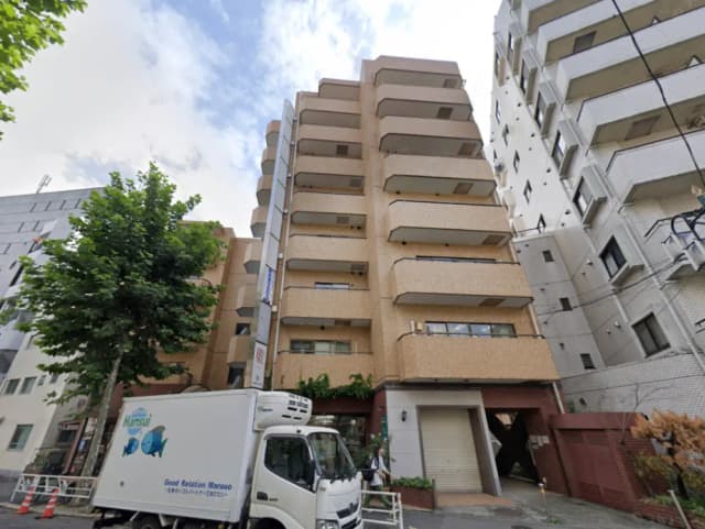 - 都市の中に立つ多層のアパートメントビル、周囲に植栽された木々と配送車が駐車している様子