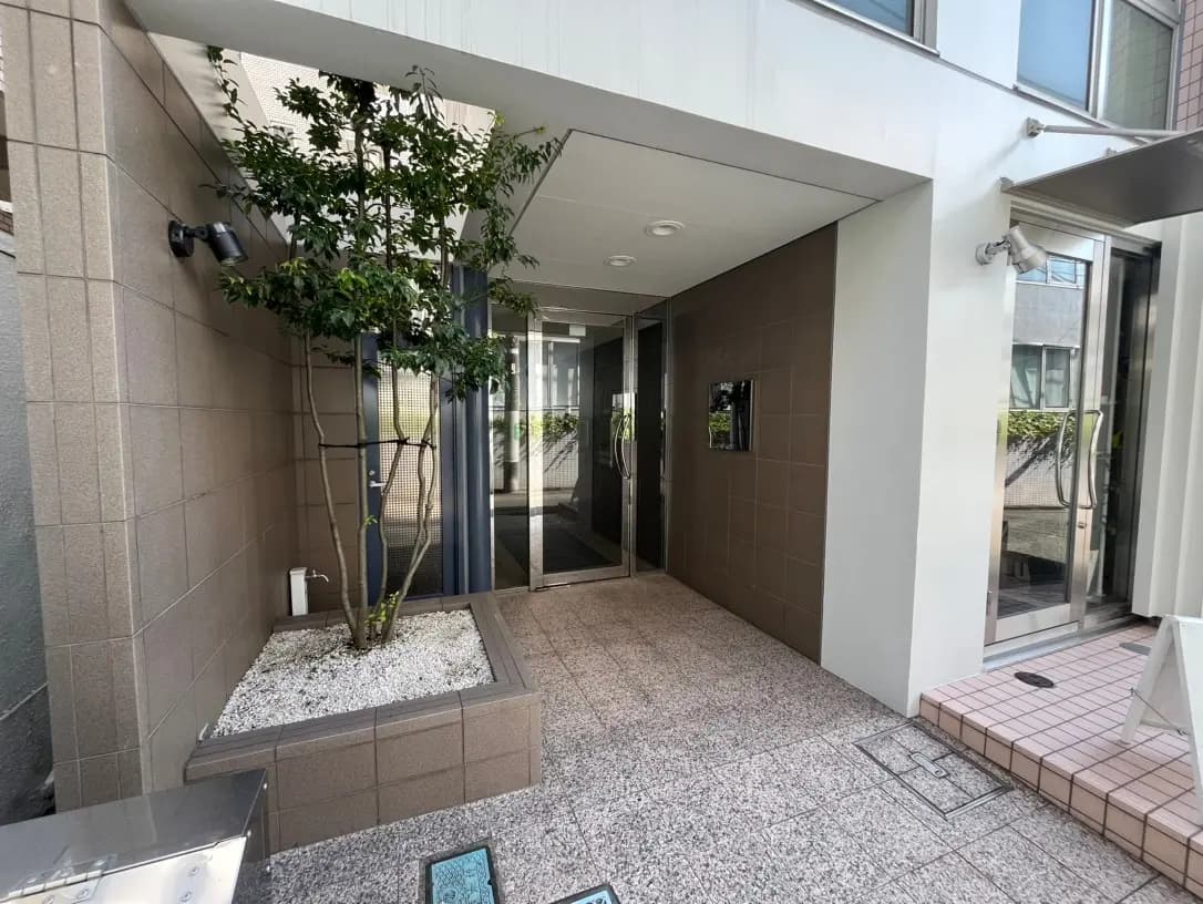 モダンな建築のエントランス、植栽された木、ガラス扉とセキュリティカメラ付きの明るい通路