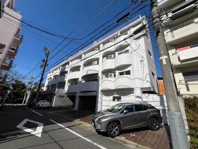 青空の下、白い外観のマンションと路上の駐車車両、電線が交差する都市景観