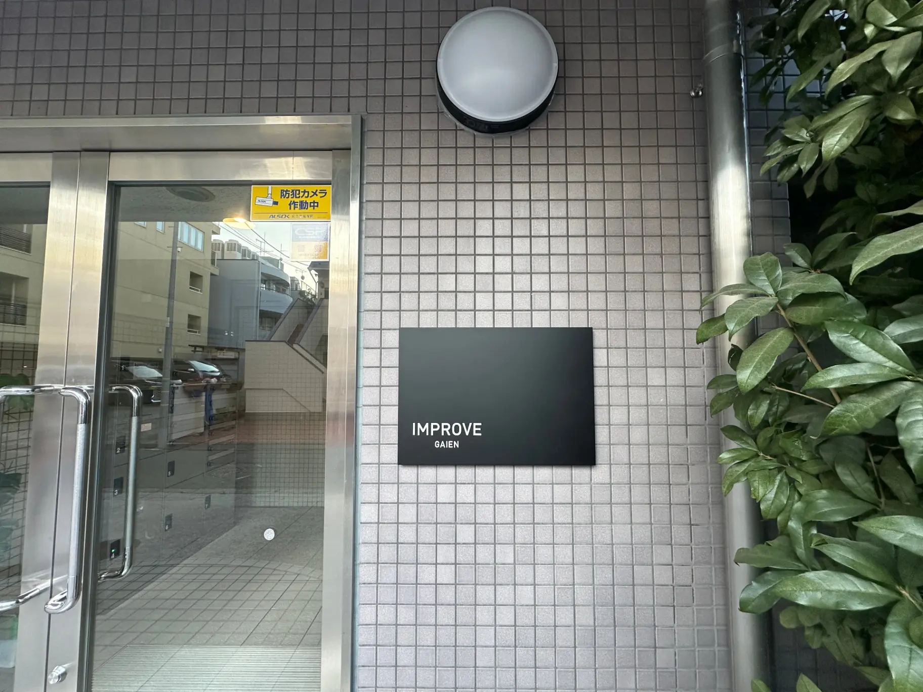 オフィスビルの入口、ガラスドアとタイル壁、外灯と植物のデコレーション、看板にはIMPROVE GAIEと表示