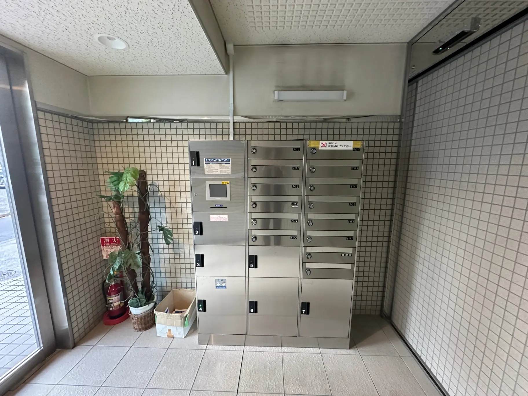 マンションのエントランスにある宅配ボックスと人工観葉植物、タイル仕上げの壁