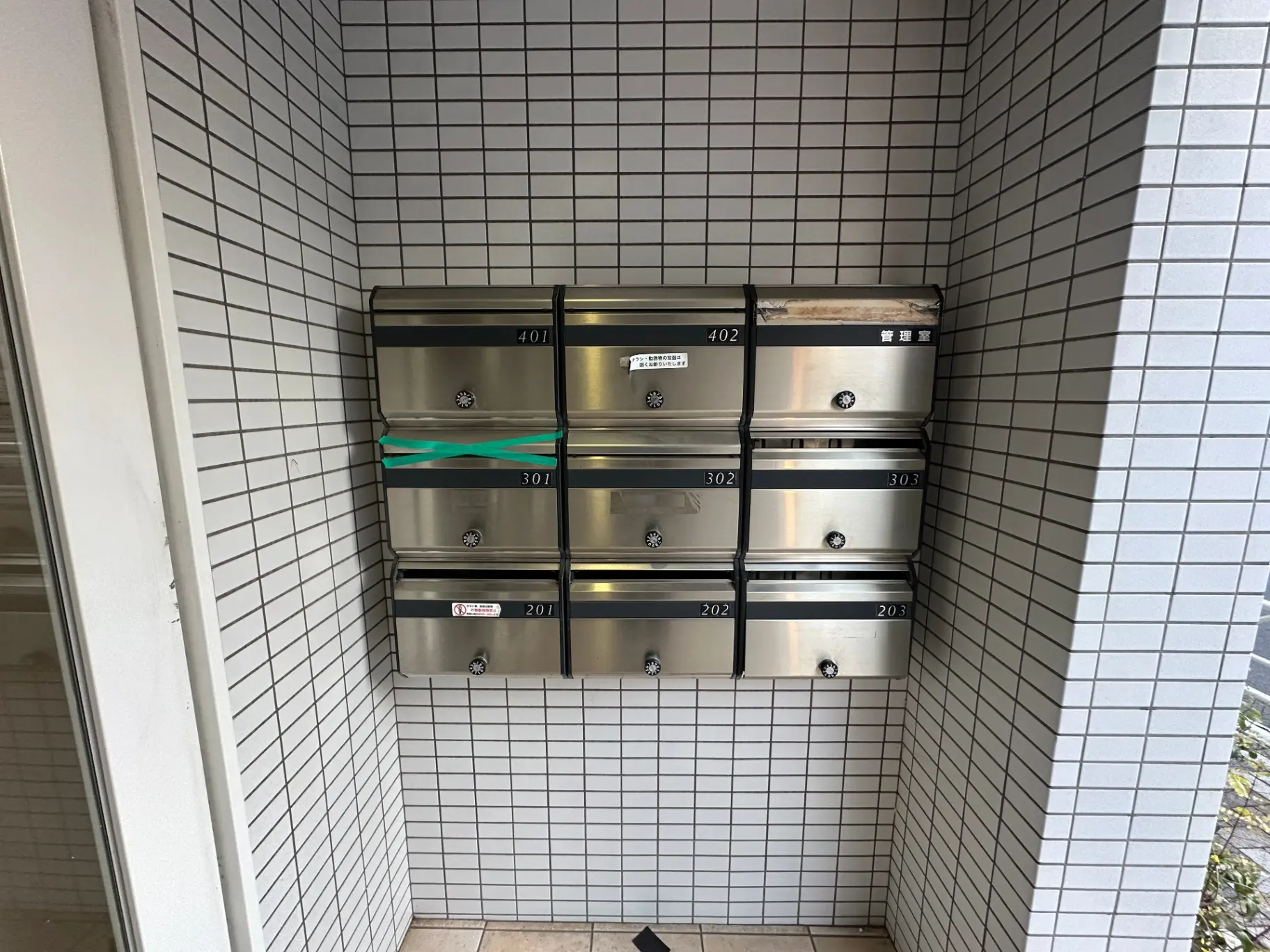 マンションの共用部にあるステンレス製の郵便受け、各戸番号が記された郵便ポスト、タイル張りの壁