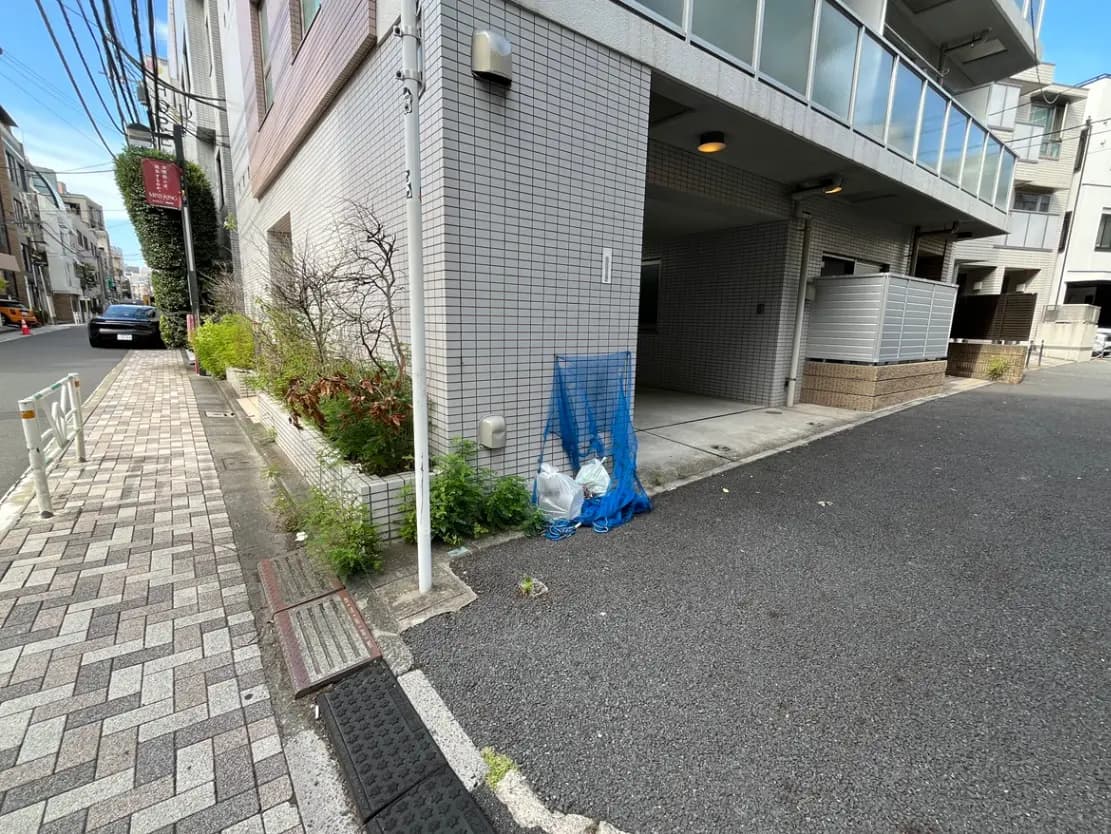 - 住宅街の街路沿い、タイル張りの建物と植栽、青いネットで包まれたゴミ袋が置かれた景観