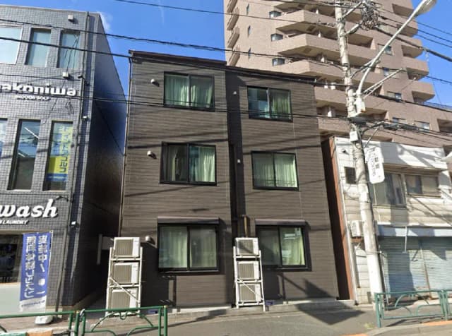 - モダンな外観のアパートメントビル、隣にはクリーニング店があり、青空の下で見える住宅街の風景