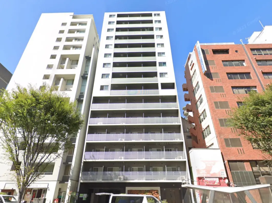 - 都市の高層マンション、白い外壁とバルコニーが特徴的、周囲には緑の木々と他の建物が並ぶ景観