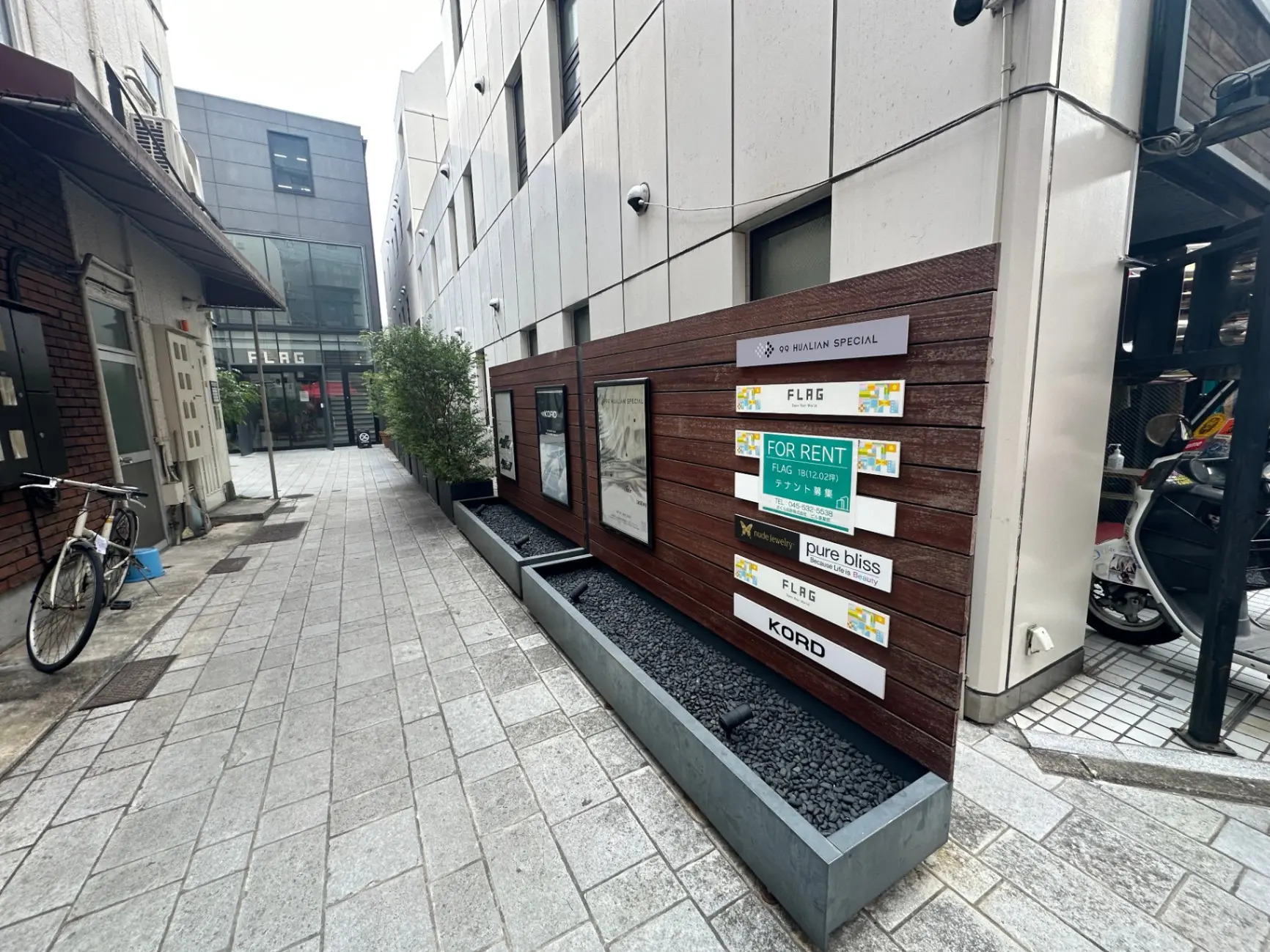 都会的な通りにあるビルの案内看板、賃貸情報が記載された看板、周囲には自転車やスクーターが見える外観