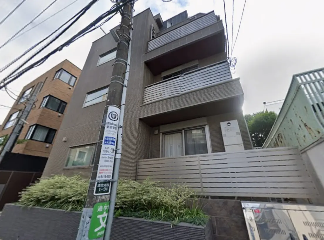- モダンなデザインの4階建てアパートメント、シンプルな外観とグリーンが施されたエントランスエリア、周囲に並ぶ建物が特徴的な街並み