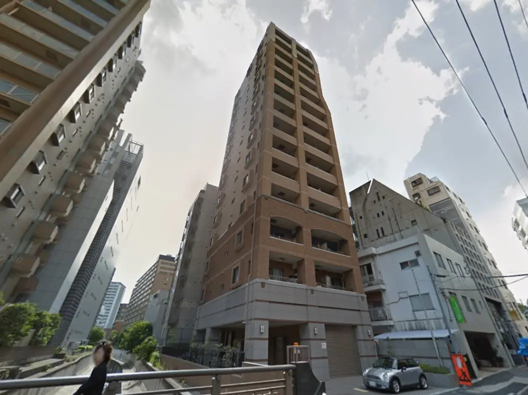 - 高層マンションの外観、周囲に並ぶビル群と青空、手前には歩道と車両が見える都市の風景