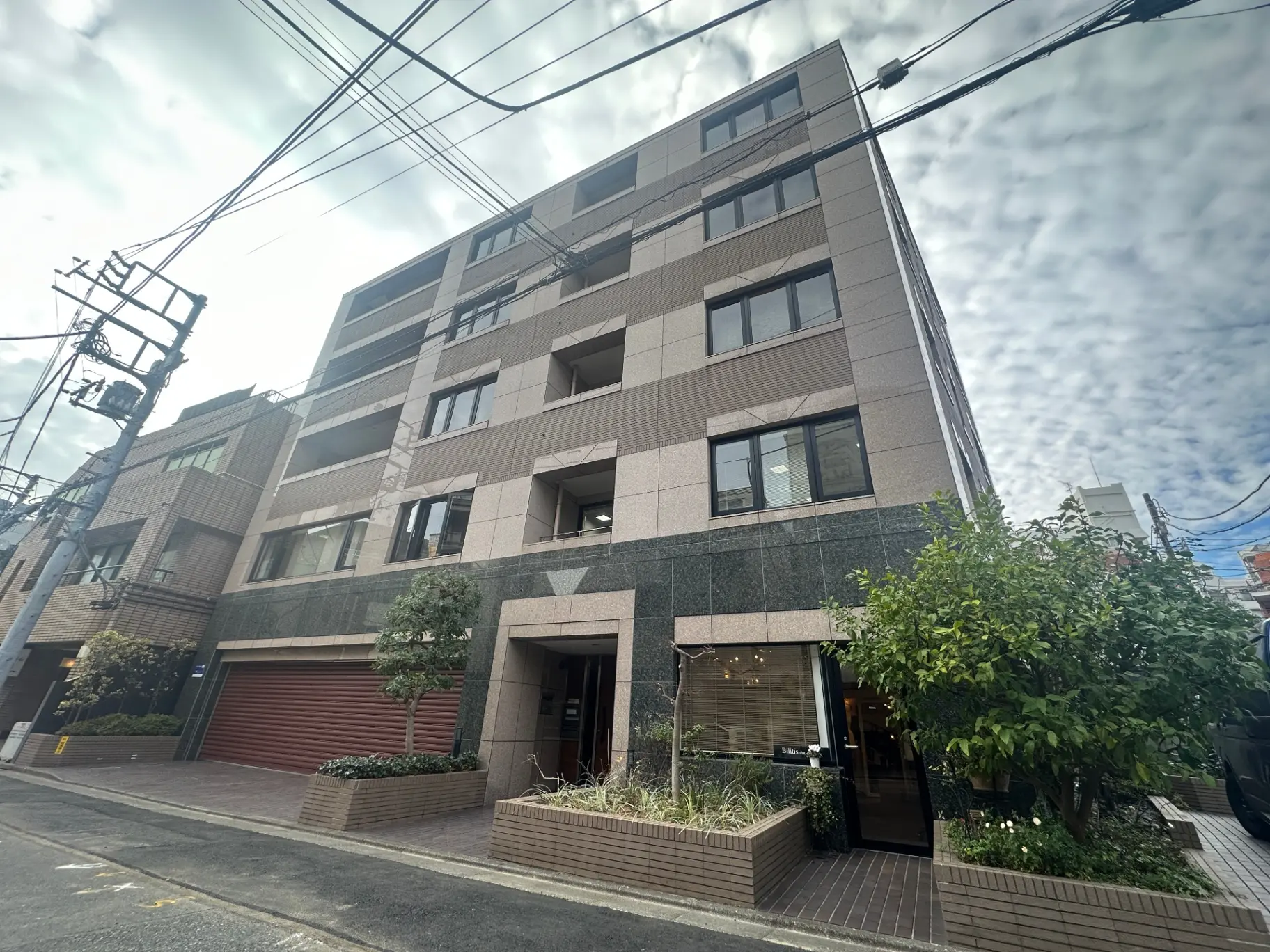 現代的な外観の都市型マンション、植栽付きのエントランス、青空と電線が交差する街並み