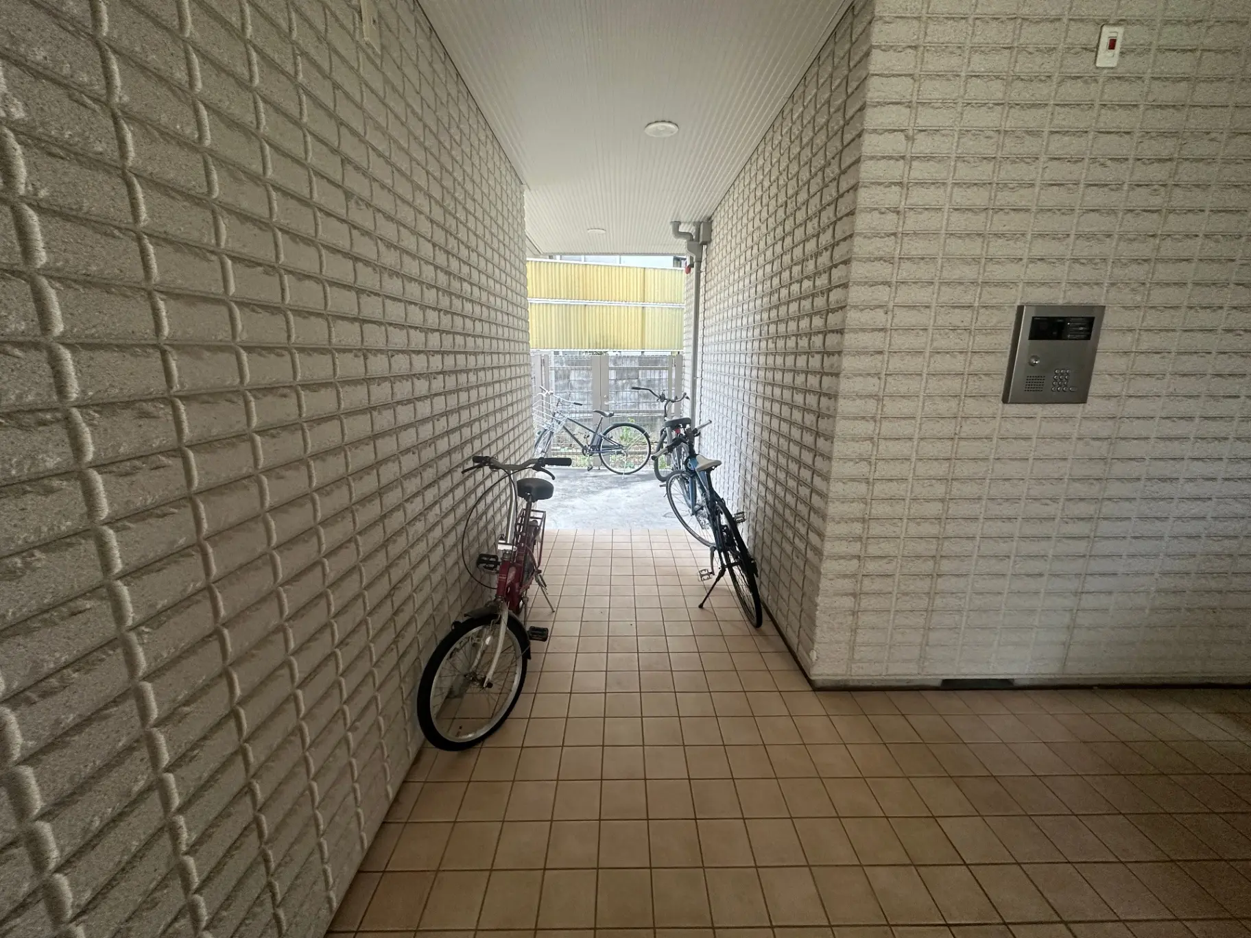 白いレンガ壁の自転車駐輪スペース、インターホンと明るい天井の照明が特徴の玄関エリア