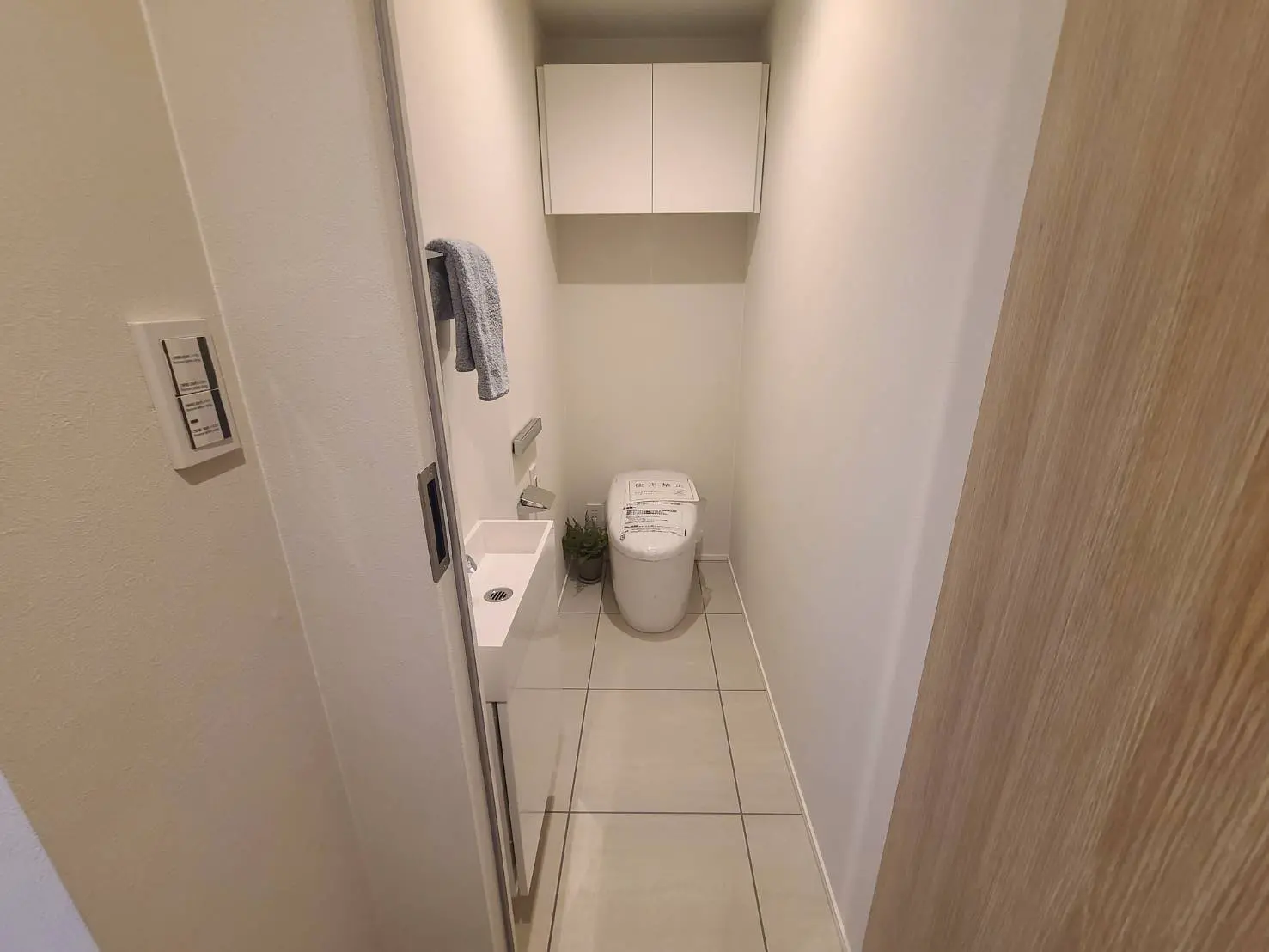 - シンプルで清潔感のあるトイレ、白い壁と床タイル、洗面台とトイレットペーパーを備えた収納スペースがあるインテリア