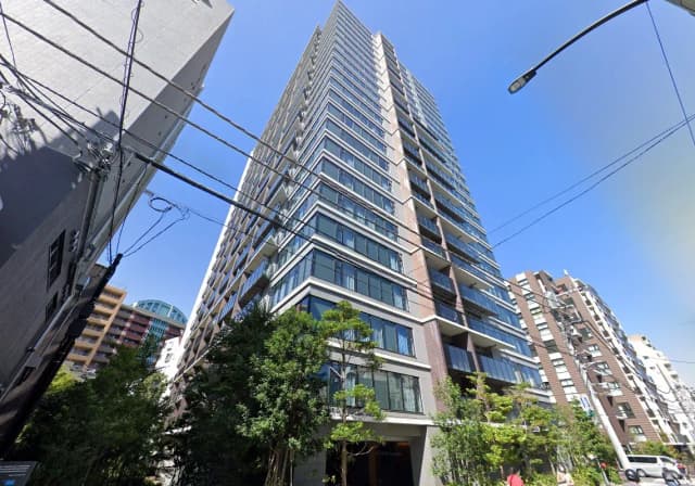 - 高層マンションの外観、周囲には住宅やビルが立ち並び、青空の下でガラス張りのバルコニーが目を引く景観