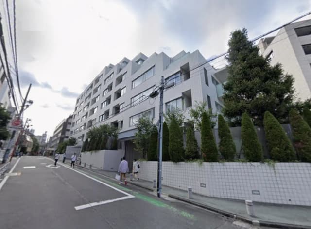 - 近代的なデザインのマンション外観、周囲に緑の植栽が施され、通りに歩行者がいる様子