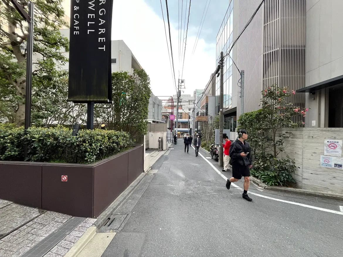 - 都会の路地にあるカフェの入り口、周囲に緑の植栽と人々が行き交う様子、遠くにはビル群が見える風景
