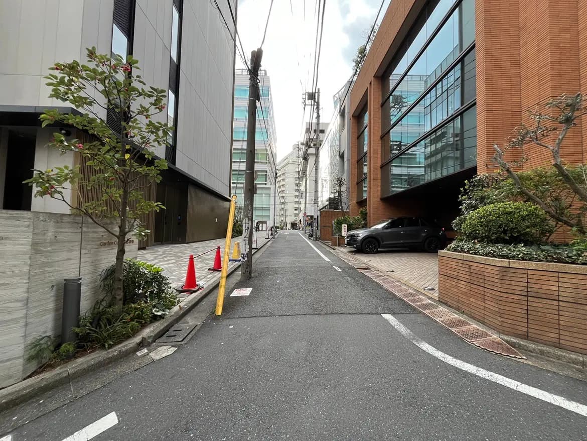 - 都市の道に沿った現代的なビルの並び、高層ビルのガラス窓と植栽が施された景観、赤いコーンが目立つ通りの風景