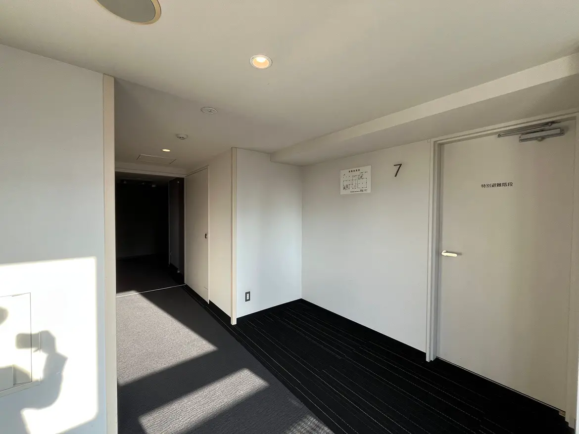 - 明るい日差しが差し込むシンプルな廊下、シックな黒いカーペットと白い壁、部屋番号7があるドアが視界に入るインテリア
