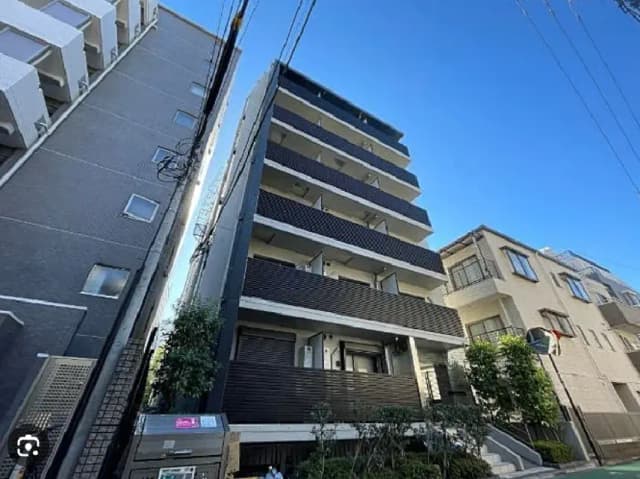 - 高層マンションの外観、青空を背景にしたデザイン性のある建物、隣接する住宅とシンプルな庭木が見える風景