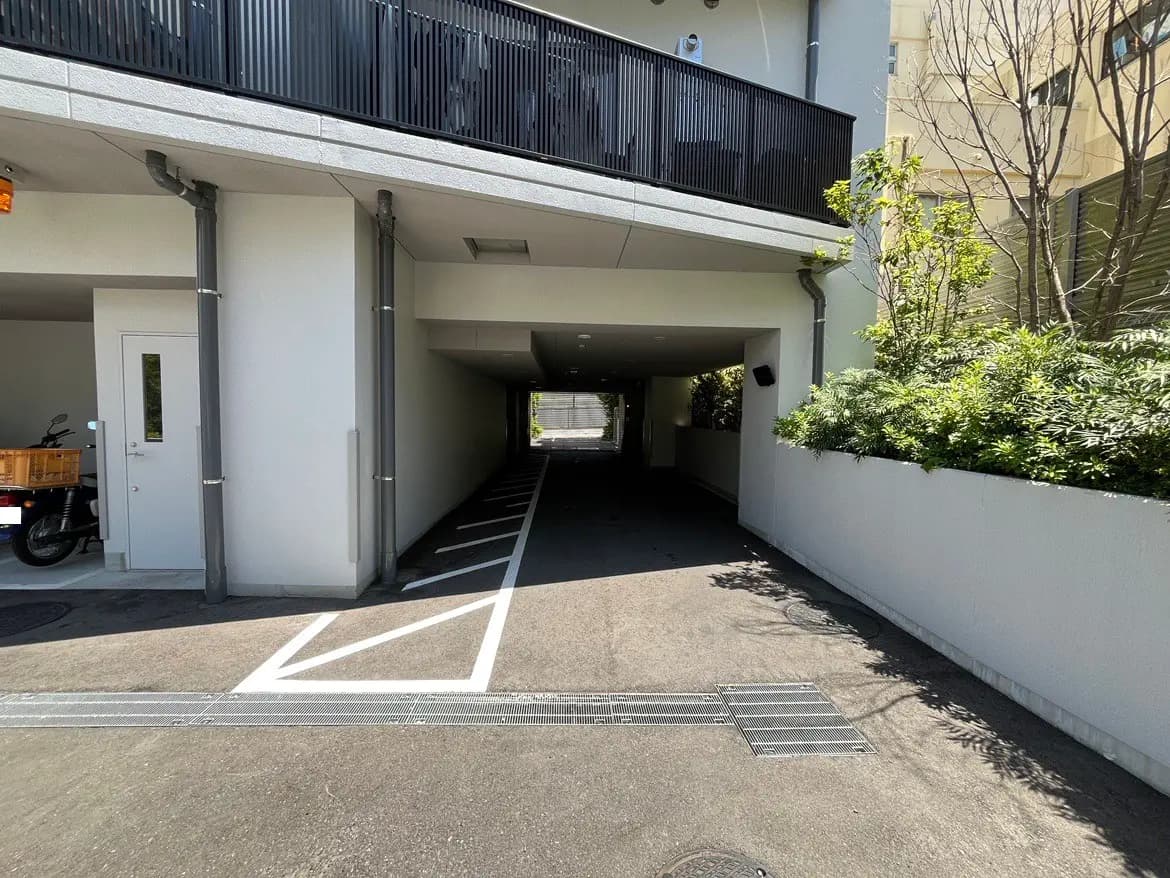 - 駐車場の出入り口、白い壁と緑の植物、軽自動車が停まるスペースのある明るい道を見渡せる風景