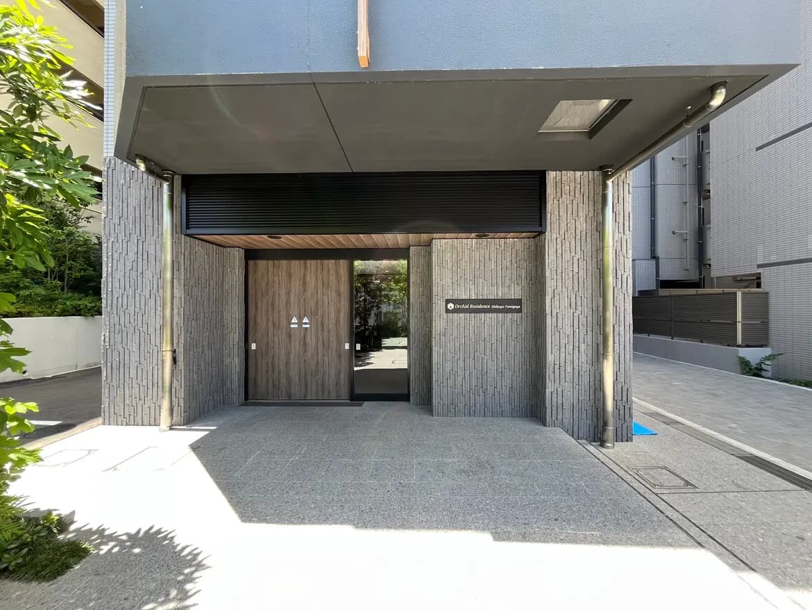 - 現代的なデザインのマンションのエントランス、木製のドアと石造りの壁、周囲に緑があふれる雰囲気