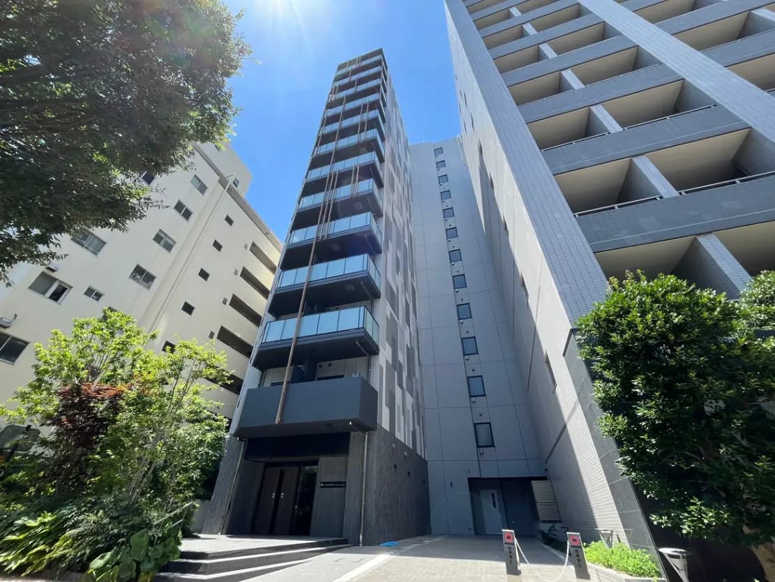 - 現代的なデザインの高層マンション、青空の下にそびえ立つ外観、周囲に緑豊かな植栽が配されたエントランス