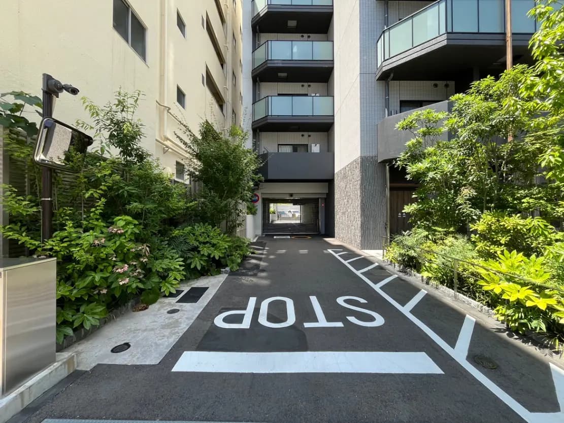 - 植栽に囲まれたマンションのエントランス、ストップサインと車両通行帯が見える道の風景